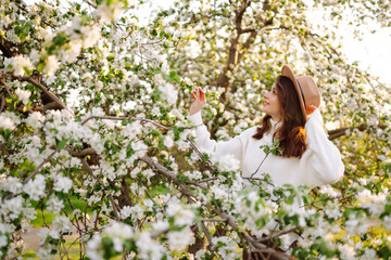 Portrait of Beautiful  Woman In hat  posing near flowering tree. Smiling young woman enjoying smell of flowers on background of spring garden.  Fashion, lifestyle. 