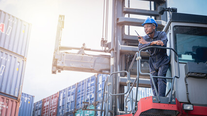 Black forklift driver in blue jumpsuit leaning on the guard rail of the forklift outdoors waiting for shipping containers to be delivered. 
