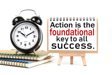 ACTION IS THE FOUNDATIONAL KEY TO ALL SUCCESS. text on white notepad paper on a stand next to our desk clock on a sideboard. on white background