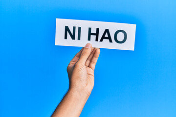 Hand of hispanic man holding chinese greeting ni hao word paper over isolated blue background.