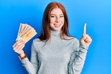 Young red head girl holding 500 norwegian krone banknotes smiling with an idea or question pointing...