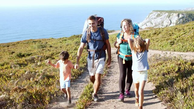Active parents with backpacks and kids walking on path at sea. Family of tourists practicing hiking, mountain trekking, enjoying vacation outdoors. Front view. Travel activity concept