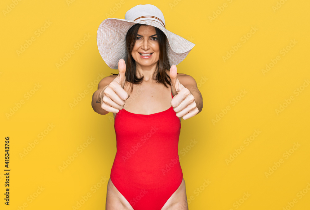 Poster beautiful brunette woman wearing swimsuit and summer hat approving doing positive gesture with hand,