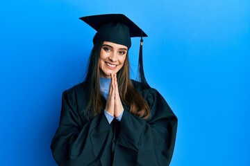 Beautiful brunette young woman wearing graduation cap and ceremony robe praying with hands together...