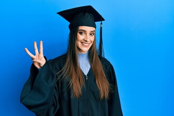 Beautiful brunette young woman wearing graduation cap and ceremony robe showing and pointing up with fingers number three while smiling confident and happy.