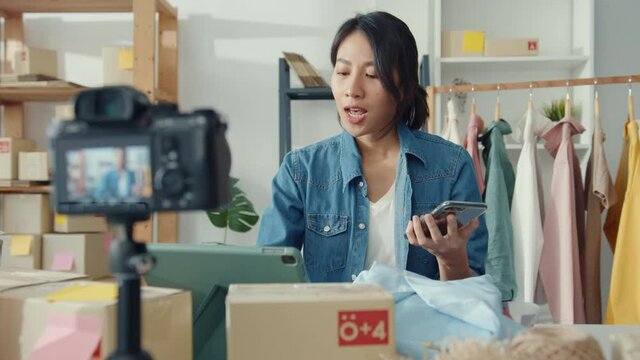 Young Asia lady fashion designer using mobile phone receiving purchase order and showing clothes recording video live streaming online at shop. Small business owner, online market delivery concept.