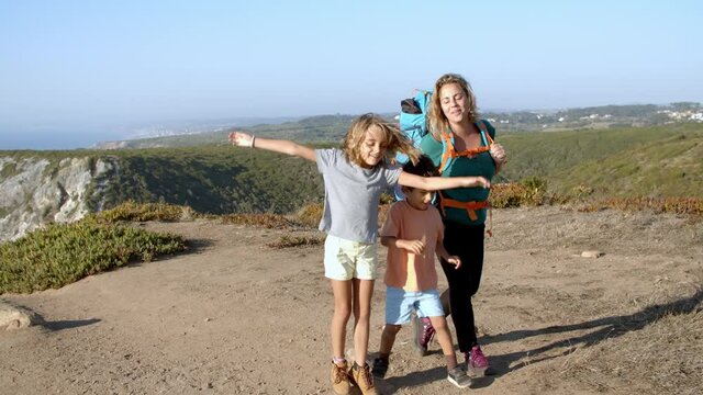 Happy kids and active mom wearing backpack, walking on mountain path, having fun. Family of active tourists practicing hiking, trekking, enjoying landscape and nature. Travel activity concept