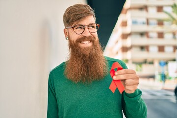 Young irish man with redhead beard smiling happy and holding red hiv ribbon leaning on the wall at...
