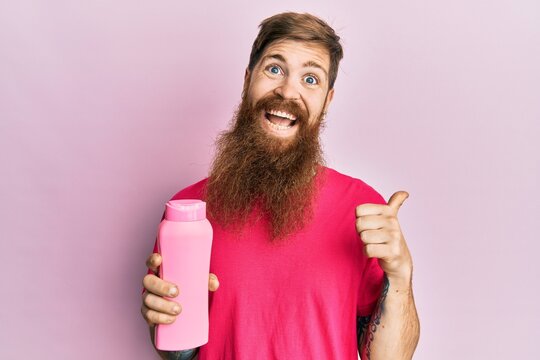 Redhead man with long beard holding shampoo bottle pointing thumb up to the side smiling happy with open mouth