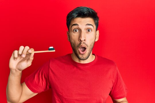 Young handsome man holding toothbrush with toothpaste scared and amazed with open mouth for surprise, disbelief face