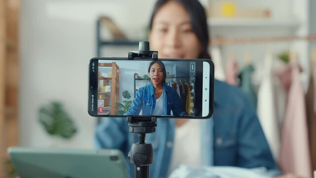 Young Asia lady fashion designer using mobile phone receiving purchase order and showing clothes recording video live streaming online at shop. Small business owner, online market delivery concept.