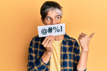 Young hispanic man covering mouth with insult message paper pointing thumb up to the side smiling happy with open mouth - obrazy, fototapety, plakaty
