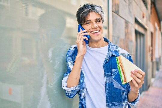 Young hispanic man smiling happy talking on the smartphone and eating sandwich at the city.