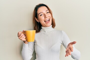 Young caucasian woman holding coffee celebrating achievement with happy smile and winner expression with raised hand