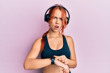 Young beautiful redhead woman wearing gym clothes, using headphones and smart watch in shock face,...