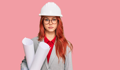 Young redhead architect woman wearing safety helmet holding blueprints thinking attitude and sober expression looking self confident