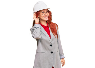 Young redhead woman wearing architect hardhat smiling with hand over ear listening an hearing to rumor or gossip. deafness concept.