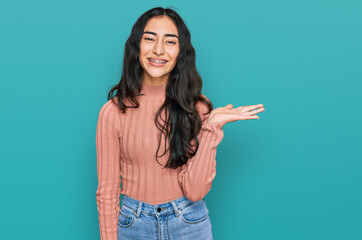 Hispanic teenager girl with dental braces wearing casual clothes smiling cheerful presenting and pointing with palm of hand looking at the camera.