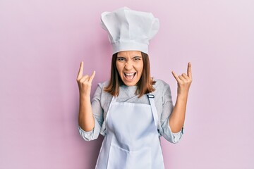 Young beautiful woman wearing professional cook uniform and hat shouting with crazy expression doing rock symbol with hands up. music star. heavy concept.