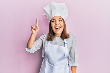 Young beautiful woman wearing professional cook uniform and hat pointing finger up with successful idea. exited and happy. number one.