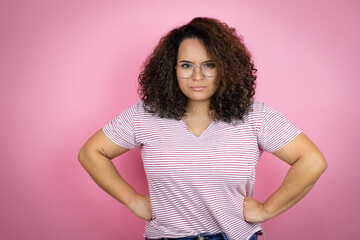 Young african american woman wearing red stripes t-shirt over pink background skeptic and nervous, disapproving expression on face with arms in waist