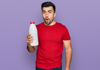 Hispanic man with beard holding liter bottle of milk scared and amazed with open mouth for surprise, disbelief face