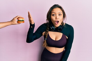 Young hispanic girl wearing sportswear rejecting burger scared and amazed with open mouth for surprise, disbelief face