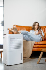 Pretty woman sitting near air purifier and moisturizer appliance near sofa monitoring air quality...