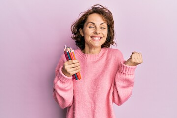 Young brunette woman holding colored pencils screaming proud, celebrating victory and success very excited with raised arm