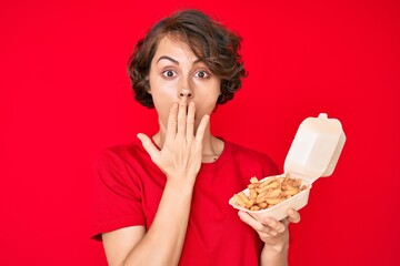 Young hispanic woman holding potato chip covering mouth with hand, shocked and afraid for mistake. surprised expression