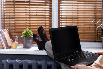 Cropped photo of a female student with her feet on the windowsill at home, holding a laptop with a black screen. Online training.