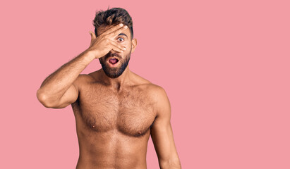 Fototapeta premium Young hispanic man wearing swimwear shirtless peeking in shock covering face and eyes with hand, looking through fingers with embarrassed expression.