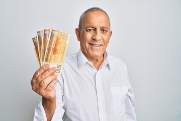 Handsome senior man holding 500 norwegian krone banknotes looking positive and happy standing and smiling with a confident smile showing teeth