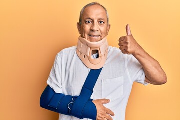 Handsome mature senior man wearing cervical collar and arm on sling doing happy thumbs up gesture with hand. approving expression looking at the camera showing success.