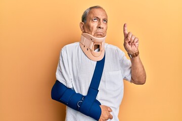 Handsome mature senior man wearing cervical collar and arm on sling pointing up looking sad and...