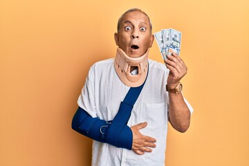 Handsome mature injured man wearing cervical collar and sling holding insurance money afraid and...