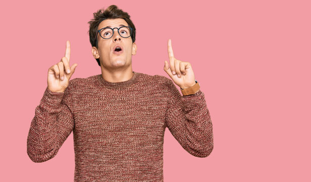 Handsome caucasian man wearing casual sweater and glasses amazed and surprised looking up and pointing with fingers and raised arms.