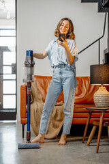 Fototapeta na wymiar Happy woman with cordless vacuum cleaning floor and using phone at home in stylish interior design of living room. Smart home concept.