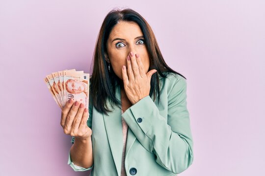 Middle age brunette woman holding 10 singapure dollars banknotes covering mouth with hand, shocked and afraid for mistake. surprised expression