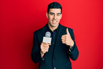 Handsome hispanic man holding reporter microphone smiling happy and positive, thumb up doing excellent and approval sign