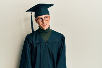 Young caucasian man wearing graduation cap and ceremony robe looking away to side with smile on face, natural expression. laughing confident.