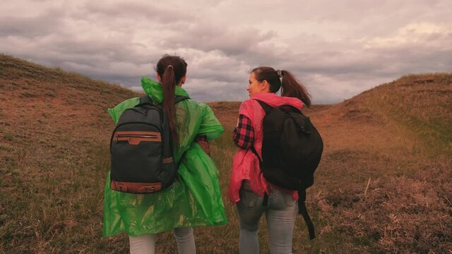 Happy young women in colored raincoats travel with backpacks in the ravine. Beautiful girls tourists go together in search of adventure. Teamwork. On day off in nature. Healthy outdoor walks