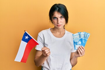 Young caucasian woman holding chile flag and chilean pesos banknotes skeptic and nervous, frowning upset because of problem. negative person.