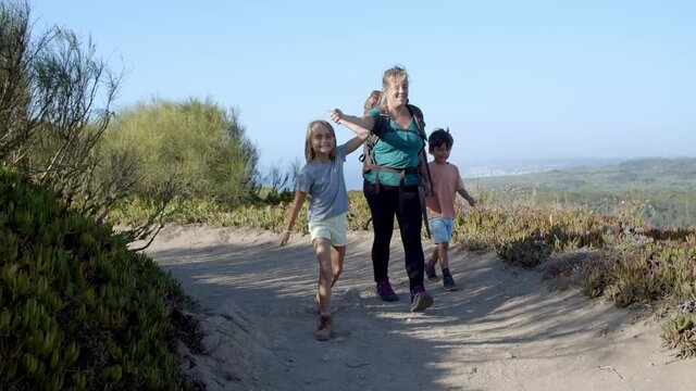 Happy kids and mom wearing backpack, walking on path in mountains, having fun. Family of active tourists practicing hiking, trekking, enjoying landscape and nature. Front view. Travel activity concept
