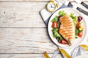 Plate of appetizing food, alarm clock and measuring tape on white wooden table, flat lay with space for text. Nutrition regime