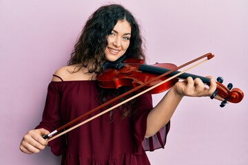 Young brunette musician woman with curly hair playing violin smiling with a happy and cool smile on...