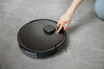 Black robotic vacuum cleaner cleaning the floor while woman sitting near sofa and relax. Smart...