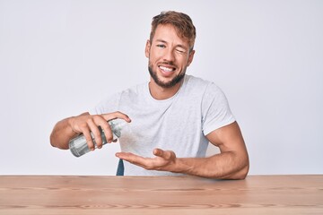 Young caucasian man using hand sanitizer gel sitting on the table winking looking at the camera with sexy expression, cheerful and happy face.