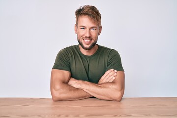 Young caucasian man wearing casual clothes sitting on the table happy face smiling with crossed...
