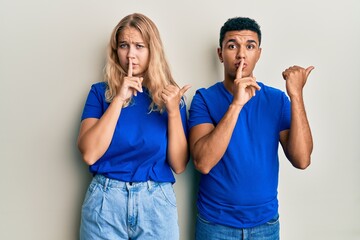 Young interracial couple wearing casual clothes asking to be quiet with finger on lips pointing with hand to the side. silence and secret concept.